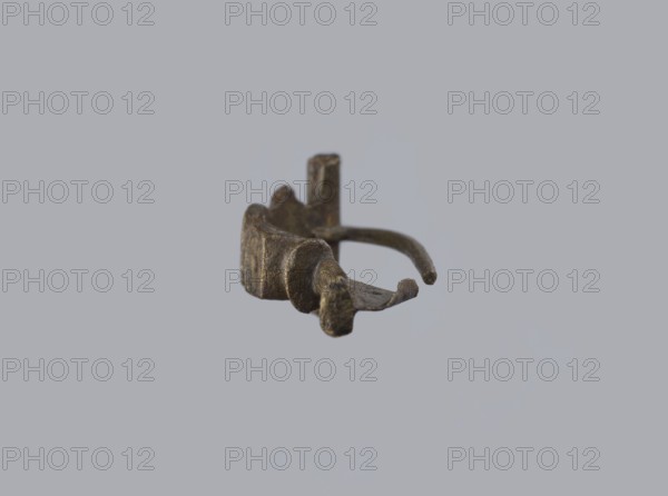 Arched fibula, unknown Roman workshop. Fibula with an arched hoop decorated with two crossbars and rectangular plates with embossed circles. The pin grows from a cylindrical crossbar. The long, flat sheath is decorated with an oval plate on the front. Preservation condition: parts of the sheath and pin are missing. Date: 75–150, Roman Empire 27 BC–476. Object type: fibula. Dimensions: height 1.6 cm, width 4.1 cm, weight 0.006 kg., A small metallic object with a rough, irregular surface and a patina of brown and dark bronze tones floats against a smooth light gray background; the object has a curved band that forms a partial loop, a few rounded protrusions and flattened extensions, and areas that appear worn or corroded in shades of brown, bronze, and hints of darker gray.