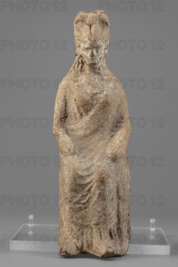 Female figurine, unknown creator/workshop, Female figure in a seated position, slightly leaning forward. The back of the figurine is not artistically developed. The woman is dressed in a robe and cloak revealing the right shoulder and falling almost to the ankles. Her hands are placed on her knees. Her hair is tied in a high bun. Traces of white paint on the figurine's head. 4th century BC, Southern Italy, figurine; height 15.4 cm, width 5 cm, depth 7.3 cm, weight 0.17 kg., A weathered seated figurine made of a beige to light brown stone with areas of off-white surface deposits, showing a draped garment and a headdress, positioned on a transparent display stand against a plain gray background.