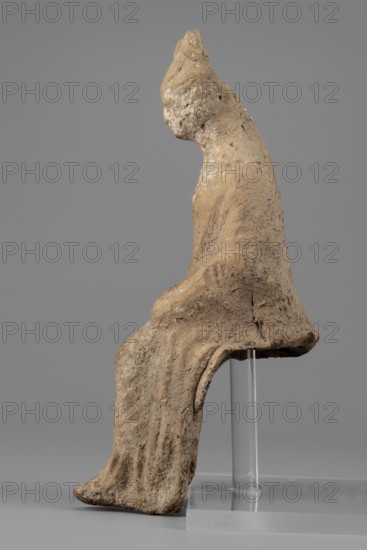 Female figurine. Female figure in a seated position, slightly leaning forward. The back of the figurine is not artistically developed. The woman is dressed in a robe and cloak revealing the right shoulder and falling almost to the ankles. Her hands are placed on her knees. Her hair is tied in a high bun. Traces of white paint on the figurine's head. 4th century BC, Southern Italy. Terracotta figurine, height 15.4 cm, width 5 cm, depth 7.3 cm, weight 0.17 kg., A weathered, rough-textured sculpted figure seated on a clear support, shown in profile with a rounded head and a draped garment that falls over the legs; the surface appears eroded with uneven edges and pitted areas. The sculpture is primarily tan and beige with spots of off-white and darker brown mottling; the support and base are clear and translucent, and the background is a uniform gray.