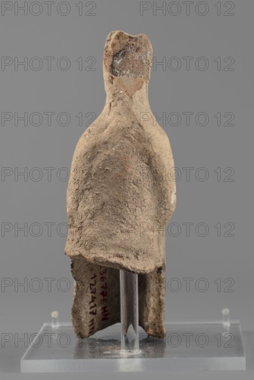 Female figurine, Female figure in a seated position, slightly leaning forward. The back of the figurine is not artistically developed. The woman is dressed in a robe and cloak revealing the right shoulder and falling almost to the ankles. Her hands are placed on her knees. Her hair is tied in a high bun. Traces of white paint on the figurine's head. 4th century BC, Southern Italy. Terracotta figurine, height 15.4 cm, width 5 cm, depth 7.3 cm, weight 0.17 kg., A worn sculptural form resembling a simplified human figure stands on a clear rectangular base with a clear vertical support; the figure has an uneven textured surface with a rough, sandy appearance and colors of beige, tan, light brown, and touches of darker brown, with small areas showing lighter whitish spots; the head is blocky and slightly darker at the top, the torso tapers downward and shows irregular surface pitting, and the lower portion is hollowed and broken, revealing the inner rough edges and an opening that exposes t...