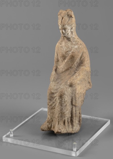 Female figurine in a seated position, slightly leaning forward. The back of the figurine is not artistically developed. The woman is dressed in a robe and cloak revealing the right shoulder and falling almost to the ankles. Her hands are placed on her knees. Her hair is tied in a high bun. Traces of white paint on the figurine's head. 4th century BC, Southern Italy. Terracotta figurine, height 15.4 cm, width 5 cm, depth 7.3 cm, weight 0.17 kg., A seated figurine made of rough textured material sits on a clear rectangular base, with a muted palette of brown and beige on the figure and a gray background; the figure wears draped clothing with visible folds and has a rounded head with subtle facial contours, and the base is translucent with a few small clear pegs at its corners.