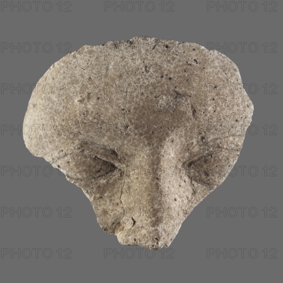Fragment of a stucco mask; upper part of a sarcophagus mask showing forehead, eyes and nose. Sarcophagus mask, stucco, height 8.8 cm, width 8.9 cm. 2nd century, Roman period, Egypt., A roughly triangular, weathered stone fragment with a textured, pitted surface appears against a flat gray background; the stone shows a palette of beige, tan, light brown, and specks of darker brown and black, with areas of lighter off-white highlights; the shape suggests shallow carved depressions resembling eye sockets and a raised central ridge resembling a nose, with irregular edges, small cracks, and rough granular detail across the surface.