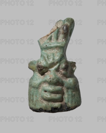 Amulet in the shape of Bes's head decorated with a crown. The grotesque face with sideburns is shaped similar to a triangle, the base of which is a flat lower part of the face. The back of the amulet is smooth, with a transverse hole at the height of the crown. 7th–4th century BC; Late Period, Egypt. Amulet; height 2.5 cm, width 1.4 cm., A small sculpted object resembling a stylized head with exaggerated facial features, shown against a plain light gray background; the surface is uneven and worn with a textured, pitted appearance and visible chips, especially near the top; colors visible include shades of green, turquoise, pale beige, brown, and small areas of dark gray or black; the form shows raised ridges and rounded protrusions suggesting eyes, a pronounced nose, and a layered mouth, with a taller irregular extension rising from the top that has both smoother and rougher areas.