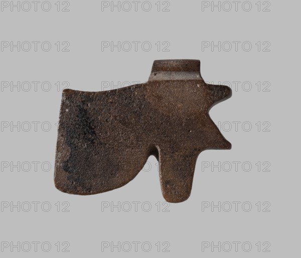 Amulet - udjat eye, udjat eye without internal modeling. Pendant on the edge of the eyebrow. 7th–4th century BC; Late Period, Egypt. Amulet. Height 2.2 cm; width 1.0 cm., A roughly shaped, flat object with an irregular silhouette resembling an abstract animal or tool is centered against a plain light gray background; the object has a rough, textured surface and appears in shades of brown, dark brown, and black with subtle lighter brown areas, showing worn edges and small pitted spots, and it features a broader rounded section on one side tapering into a narrower protruding part with a small notch between them.