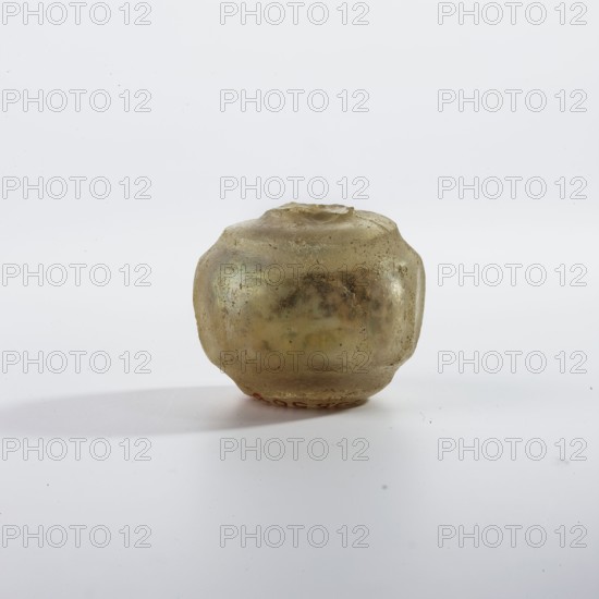 Bottle, Islamic art, Persian, bottle decorated with a row of oval facets and engraved circles separating the collar and foot; yellow-green glass with wheel-cut decoration; 9th–11th century; Iran; vessel; height 2.6 cm, width 3.1 cm, A small rounded object with an irregular, slightly flattened top and bottom sits centered on a plain surface, appearing translucent with a weathered, pitted texture; its colors include pale yellow, muted green, and hints of brown, and it casts a soft shadow to the left on a mostly white background.