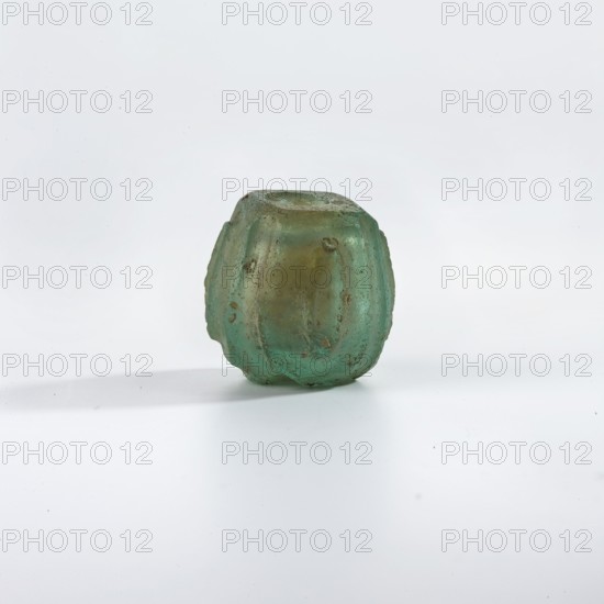 Bottle, Islamic, Persian; blue-green glass bottle decorated with small almond-shaped ovals in an oval field bounded by grooves extending to the edge and intersecting there; wheel-cut decoration. 9th–11th century. Origin: Syria, or Iran . Vessel; height 2.7 cm, diameter 2.8 cm., A small rounded object with a ridged, lobed surface sits on a flat pale background, showing a translucent green color with areas of brownish discoloration and rough texture; the object casts a soft gray shadow and appears worn with irregular surface marks and small chips.