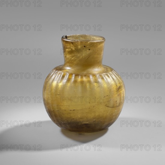 Bottle, household glass vessel, decorated with fluting on the shoulders that flow into a wide, low neck; slightly concave bottom; green-yellow glass with bubbles; Abbasid dynasty, 9th–10th century; Iran; Persian; height 8 cm, diameter 6.7 cm., A rounded glass vessel with a short neck and slightly flared rim sits against a neutral background; the glass is translucent amber and yellow with variations in tone and some darker brown areas, showing surface irregularities and faint streaks. The body has a series of vertical rib-like grooves near the shoulder and a smooth lower portion that reflects light, producing subtle highlights and soft shadow beneath the vessel. The neck appears somewhat uneven at the rim, and small bubbles or inclusions are visible within the glass.