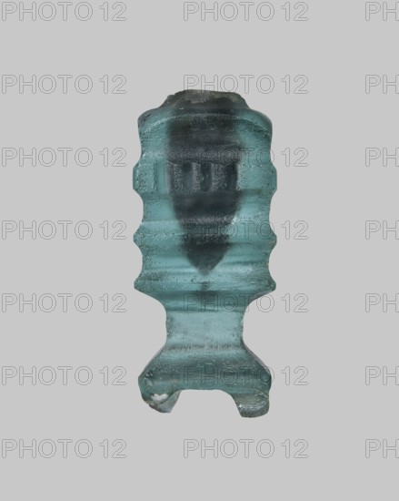 Cosmetic bottle, ; Islamic, Arabic culture; 9th–11th century, Egypt. Thick-walled blue glass vessel, height 4.1 cm, width 1.7 cm. On the body one convex band decorated with fine horizontal grooves; flat arms; heavily carved base with four feet; wheel-cut decoration., A single translucent object with a textured surface centered against a plain light gray background, showing shades of aqua, teal, and darker green-blue; the object has an irregular vertical shape with a rounded top, constricted midsections creating wavy contours, and a broader lower portion that forms a short pedestal with two small protrusions at the base; surface details include visible ridges, smooth and rough patches, and variations in translucency producing darker internal areas and lighter outer highlights.
