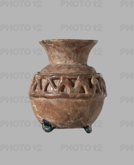 Cosmetic bottle. Spherical body decorated at the top with a zigzag pattern of applied glass thread. Short, flared neck. Colorless glass bottle with blue glass feet and applied decoration. 7th–8th century, possibly Egypt or Syria. Height 4.6 cm; diameter 4 cm. Islamic/Arabic cultural context., A small ceramic vessel with a rounded body and a flared neck sits against a plain gray background; the surface of the vessel is textured and worn, showing various shades of brown and tan with lighter and darker patches, and a band of open triangular cutouts encircles the upper body, forming a decorative pattern; the rim has a small chip, and the vessel rests on a few small rounded feet that display green and dark bluish tones.