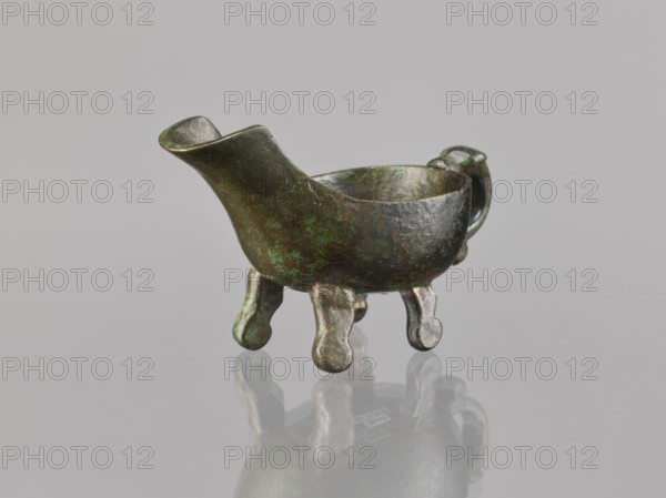 Yi-type vessel, Chinese; Chinese art; ceremonial/ritual vessel; Qing dynasty 1644–1911; vessel on four legs one broken off; underside: image of a human; 19th century, China; cult object; height 6.4 cm, width 11.0 cm, depth 4.5 cm., A small metal vessel with a spout and handle sits on a reflective surface; the vessel has a rounded bowl supported by a group of short, rounded legs and shows a pitted, aged texture. The object displays mottled colors of dark green, brown, and hints of black with areas of brighter green patina and some lighter metallic tones on the legs. The background is a uniform light gray, and the vessel casts a soft reflection beneath it.
