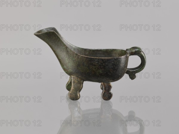 Yi-type vessel, Chinese, Chinese art, ceremonial/ritual vessel, Qing dynasty 1644–1911, 19th century, China, cult object; vessel on four legs one broken off; underneath, an image of a human; height 6.4 cm, width 11.0 cm, depth 4.5 cm., An aged metal vessel with a dark green and brown patina sits on a reflective surface; it has a long spout on one end, a rounded handle on the opposite end, a shallow open bowl body, and a few short, rounded legs that lift it slightly, with visible mottled textures and variations of green, brown, and hints of black on its surface.