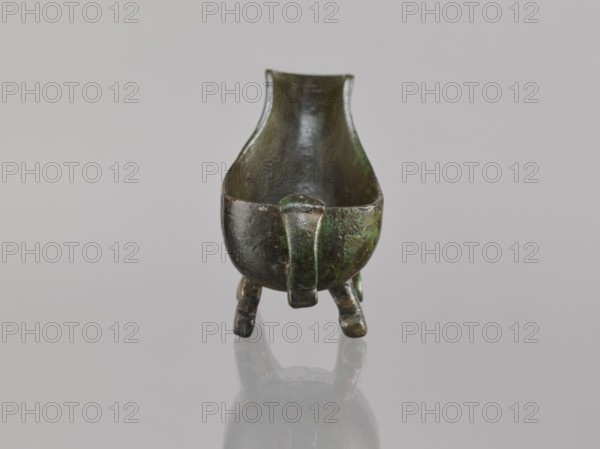 Yi-type vessel, Chinese, ceremonial ritual vessel, Qing dynasty 1644–1911, vessel on four legs one broken off with an image of a human underneath, cult object, 19th century, China, height 6.4 cm, width 11.0 cm, depth 4.5 cm, A small, boat-shaped metal vessel with a dark patina and areas of green discoloration sits centered on a smooth, light gray surface; the vessel has a rounded body, a raised rim that tapers toward the back, a short vertical handle-like projection at the front, and a group of short, curved legs supporting it, with reflections visible beneath it.