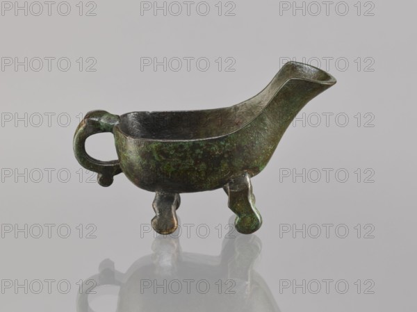 Yi-type vessel, Chinese, Qing 1644–1911, 19th century, ceremonial ritual vessel, vessel on four legs one broken off; underneath an image of a human, cult object; height 6.4 cm, width 11.0 cm, depth 4.5 cm, An aged metal vessel with a patinated surface in shades of green, dark brown, and hints of black, shaped like a small boat with a curved spout at one end and a rounded handle at the other; the body rests on a few short, rounded legs and the interior appears darker with a rough texture, while the exterior shows mottled green corrosion and darker areas, all sitting on a reflective gray surface that mirrors the object faintly.