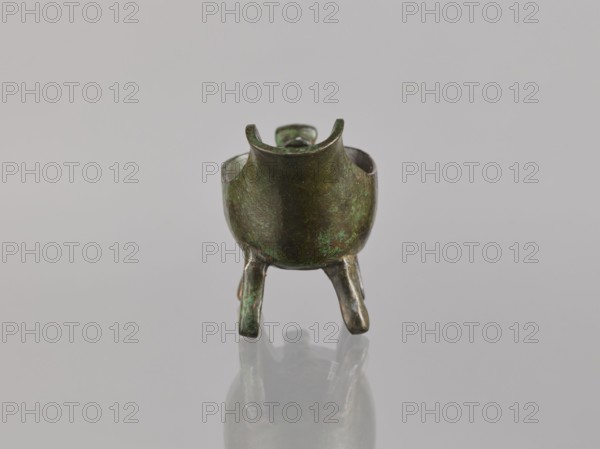 Yi-type vessel, Chinese, Qing 1644–1911, 19th century, China. Ceremonial/ritual vessel on four legs one broken off; underneath, an image of a human. Cult object. Materials/technique not specified. Dimensions: height 6.4 cm, width 11.0 cm, depth 4.5 cm., A small metallic tripod vessel with a rounded body and three short legs stands on a reflective surface; the vessel shows a mottled green and brown patina with darker and lighter areas, and the interior edges and rim are visible with similar coloration, all set against a plain light gray background.