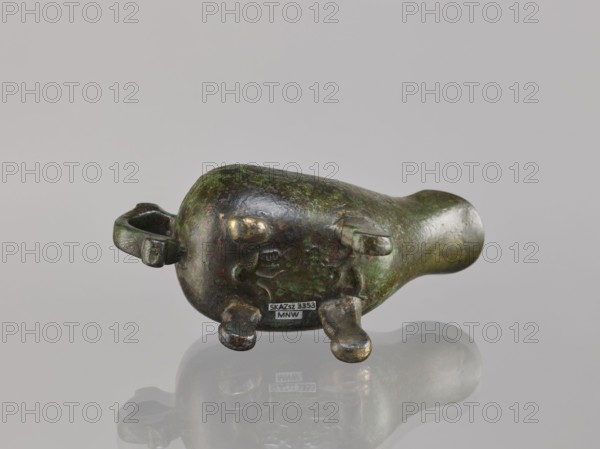 Yi-type vessel, Chinese culture, Chinese art, ceremonial ritual vessel, Qing dynasty 1644–1911, 19th century, China, vessel on four legs one broken off; underneath, an image of a human, cult object, height 6.4 cm, width 11.0 cm, depth 4.5 cm., A small rounded sculptural object rests on a reflective surface, showing a patinated finish with visible green and dark brown areas and spots of brighter bronze where the surface is worn; it has a bulbous body tapering into a narrower rounded end, with a looped handle on one side and several short stumpy protrusions on the underside; the backdrop is a smooth neutral gray, and the object casts a soft reflected silhouette on the surface beneath it.