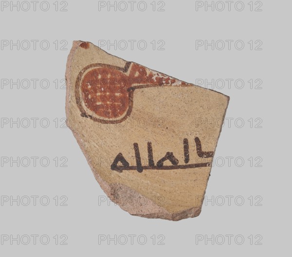 Fragment of a bowl bottom with an Arabic pseudo-inscription, decorated with an inscribed Arabic motif and a fragment of an arabesque motif after G. Pierrat. 8th–10th century, Edfu, Egypt. Ceramic fragment; height 8.1 cm, width 5.9 cm, depth 0.9 cm., A broken pottery shard with a rough, beige-tan surface showing painted decoration including a round, reddish-brown circular motif with a darker brown outline and a few pale spots, and a darker brown linear design below; the shard edges are irregular and show exposed pinkish-brown clay, set against a light gray background.