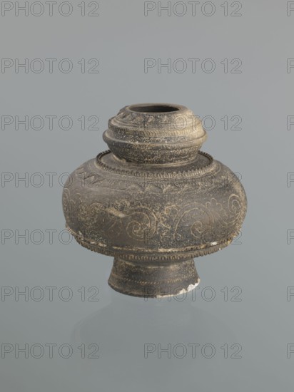 Jug with a bulging belly on a high foot. Ceramic jug decorated with plant twig motifs and raised geometric and geometric-floral patterns. Persian/Islamic style, Central Asia, 16th–18th century, height 18.7 cm, width 18.3 cm., An aged round ceramic vessel with a short narrow neck and a flared foot, shown against a plain gray background, displaying a textured surface with carved swirling decorative patterns and bands of raised ridges; the object appears worn with areas of lighter abrasion and small chips revealing a paler material beneath. Colors visible include dark gray, muted brown, light beige, and off-white.