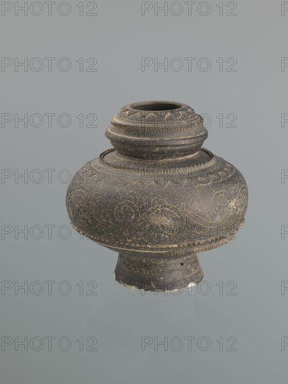 Jug, Persian, 16th–18th century, Central Asia, earthenware vessel with bulging belly on a high foot, height 18.7 cm, width 18.3 cm. Decoration: plant twig and raised geometric and geometric-floral ornament., An ornate round vessel with a flared base and a short neck, shown against a plain gray background; the object appears dark brown to black with lighter worn areas and fine incised decorative patterns of swirling lines and leaf-like motifs around its bulbous body, and textured horizontal bands and ridges around the neck and shoulder.