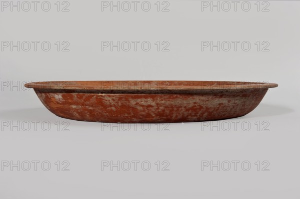 Terra sigillata plate. Plate with a flat bottom and almost vertical side walls. Narrow, flat rim. Dark orange glaze. 1st half of the 3rd century. Tunisia. Plate, diameter 36.5 cm, height 5.1 cm., A wide shallow round dish with a low rim sits centered against a pale background; the dish has a weathered, mottled surface in shades of reddish-brown and muted gray with areas of lighter beige wear, and the scene also shows pale gray and off-white tones in the flat surface and background.
