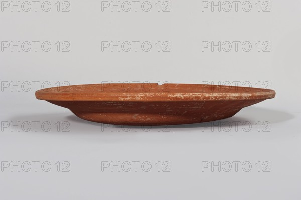 Terra sigillata plate. Flat plate with a very low foot and a rather flat, wide rim. Made of clay with orange glaze. 2nd half of the 4th century. Found in Tunisia. Plate, diameter 33.5 cm, height 5.6 cm., A low, shallow terracotta dish with a wide, flat rim and a slightly raised base sits centered against a pale background; the dish shows a weathered surface with irregular pale patches and scratches, and its colors include reddish-brown, lighter beige scuffs, and subtle gray shadows on the underside and background.