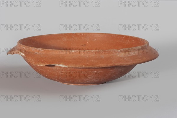 Terra sigillata plate, 2nd half of the 5th century, Tunisia; plate; height 17.4 cm, width 4.6 cm., A shallow terracotta bowl with a wide flanged rim sits against a plain light gray background; the bowl is a warm reddish-brown color with areas of lighter dusty residue and small worn patches, showing a rough matte surface and a few chips along the rim and one chipped lip that exposes a paler clay interior, and there is a subtle shadow beneath the bowl on the gray surface.