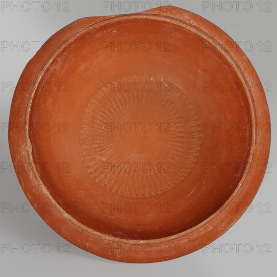 Terra sigillata plate, 2nd half of the 5th century, Tunisia; plate, height 17.4 cm, width 4.6 cm., A round shallow bowl viewed from above, made of terracotta with a matte surface in shades of reddish-brown and orange, with a slightly raised rim showing lighter worn edges and some small scuffs, and a decorative circular pattern of radiating ridges pressed into the center in similar reddish-brown tones against a neutral gray background.