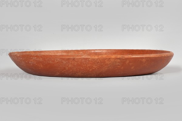 Terra sigillata plate, Roman, 325–400/420 CE, found in Tunisia; plate, height 24.9 cm, width 3.3 cm., A shallow, round clay dish with a smooth rim sits centered against a neutral background; the dish is terracotta in color with visible variations in tone and surface texture, showing a few lighter scuffs and worn areas. The background and surface beneath the dish are pale gray, and soft lighting creates subtle shadows along the dish’s lower edge.
