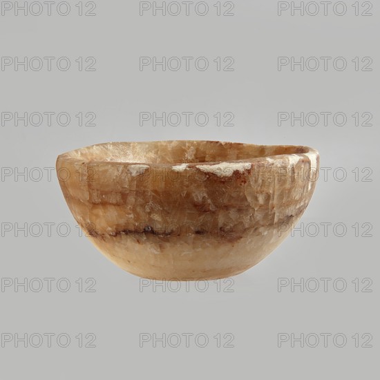 Bowl made of striped, coarse-crystalline Egyptian alabaster, waxed, with a flat bottom and gently sloping walls. 20th century, Egypt. Forgery. Vessel. Height 4 cm, width 9 cm., A shallow bowl with an irregular rim, appearing carved from stone with a textured, crystalline surface, showing a mix of warm beige, amber, light brown, cream, and hints of darker brown; the bowl sits alone against a flat light gray background.