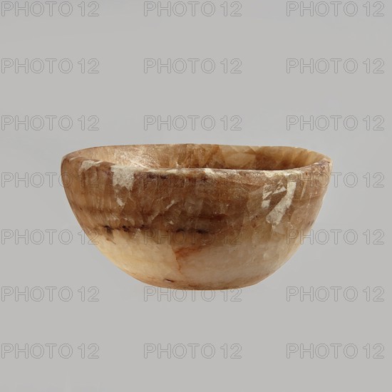 Bowl made of striped, coarse-crystalline Egyptian alabaster, waxed, with a flat bottom and gently sloping walls. 20th century, Egypt. Forgery. Vessel. Height 4 cm, width 9 cm., A rounded bowl with a smooth, slightly irregular rim sits against a plain background; the bowl's surface shows a marbled pattern of warm beige, light brown, creamy white, and hints of darker brown, with visible translucent and opaque patches and subtle variations in texture and sheen.