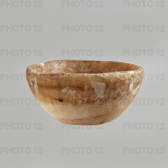 Bowl made of striped, coarse-crystalline Egyptian alabaster, waxed, with a flat bottom and gently sloping walls. 20th century, Egypt. Forgery. Vessel. Height 4 cm, width 9 cm., A shallow bowl with a rounded form made of a translucent stone showing a mix of warm beige, amber, cream, and pale brown tones, with a mottled, crystalline surface texture and irregular lighter patches and darker streaks, photographed against a smooth light gray background.