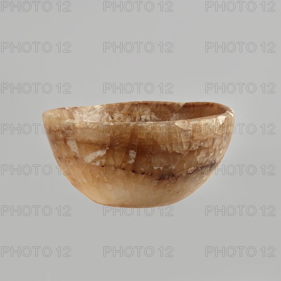 Bowl made of striped, coarse-crystalline Egyptian alabaster, waxed, with a flat bottom and gently sloping walls. 20th century, Egypt. Forgery. Vessel. Height 4 cm; width 9 cm., A shallow rounded bowl with a textured, crystalline surface in shades of brown, tan, beige, and cream, showing a mix of lighter and darker patches and subtle striations, set against a plain light gray background.