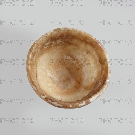 Bowl made of striped, coarse-crystalline Egyptian alabaster, waxed, with a flat bottom and gently sloping walls. 20th century, Egypt. Forgery. Vessel. Height 4 cm, width 9 cm., A roughly circular, shallow bowl with a textured, crystalline surface in shades of beige, tan, and light brown sits against a pale gray background; the bowl shows mottled patterns and translucent areas with a slightly glossy sheen and an irregular rim with small lighter patches.