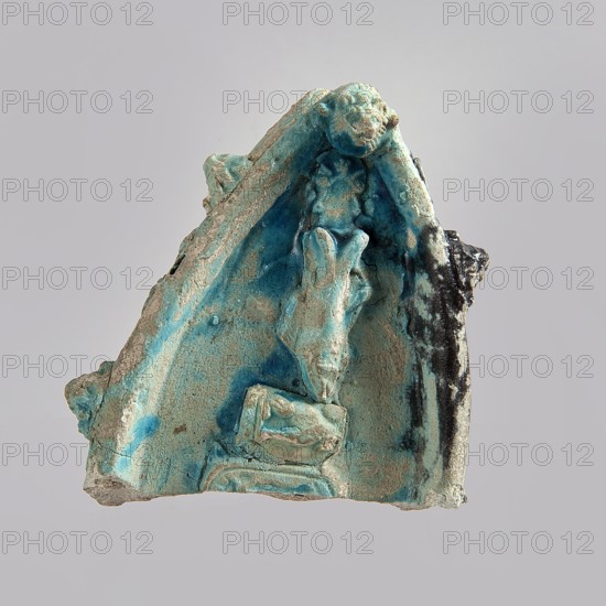Fragment of a votive boat, votive offering; prowl or stern of a boat with decoration in the form of glued appliqués. On the sides from the top: four lion heads. On the deck: palmette, fish, sitting cat with a disc above its back, cartouche half preserved. On the outside sides: two fish, four lion heads, four rosettes, three cats with a disc, two falcons with a uraeus behind their backs, four falcons with a neb symbol and a nechacha whip behind them one partially preserved, two en face faces, two rosettes one partially preserved. The bottom of the boat is smooth, slightly convex. Blue glaze, discolored black in places., glazed faience. Date: c. 30 BC–395 AD, Roman period Egypt. Function: votive offering. Dimensions: height 4.5 cm, width 11 cm, depth 8.9 cm., A triangular fragment of a sculpted object with a textured, weathered surface in shades of turquoise, teal, blue, sandy beige, and areas of dark gray to black. The fragment shows a recessed central area with raised, irregular forms ...