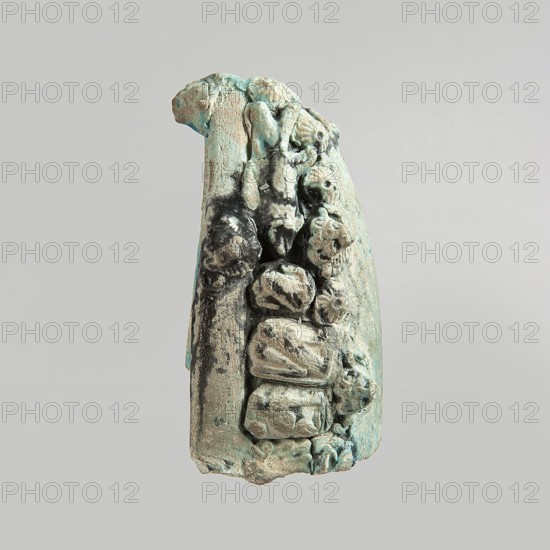 Fragment of a votive boat. Prow or stern with decoration in the form of glued appliqués. On the sides from the top: four lion heads. On the deck: palmette, fish, sitting cat with a disc above its back, cartouche half preserved. On the outside sides: two fish, four lion heads, four rosettes, three cats with a disc, two falcons with a uraeus behind their backs, four falcons with a neb symbol and a nechacha whip behind them one partially preserved, two en face faces, two rosettes one partially preserved. Bottom smooth, slightly convex. Blue glaze, discolored black in places. c. 30 BC–395 AD, Roman period, Egypt. Votive offering. Height 4.5 cm; width 11 cm; depth 8.9 cm., A vertical fragment of a textured object with an uneven, rough surface and irregular shapes arranged along one side, showing a worn, layered appearance; the visible colors include pale turquoise, light blue-green, sandy beige, muted gray, and some darker grayish-black areas, all set against a plain light gray background.