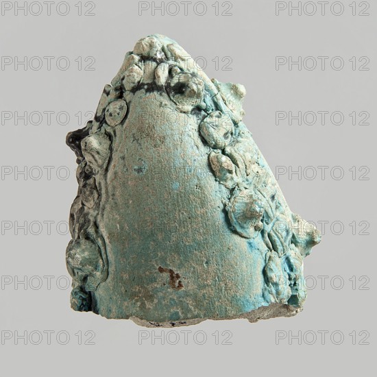 Fragment of a votive boat. Prow or stern with decoration in the form of glued appliqués. On the sides from the top: four lion heads. On the deck: palmette, fish, sitting cat with a disc above its back, cartouche half preserved. On the outside sides: two fish, four lion heads, four rosettes, three cats with a disc, two falcons with a uraeus behind their backs, four falcons with a neb symbol and a nechacha whip behind them one partially preserved, two en face faces, two rosettes one partially preserved. Bottom smooth, slightly convex. Blue glaze, discolored black in places. c. 30 BC–395 AD, Roman period, Egypt. Votive offering. Height 4.5 cm; width 11 cm; depth 8.9 cm., An irregular, cone-shaped object with a rough, textured surface covered in a patina of turquoise and pale green, with areas of darker blue-green and small patches of brown and beige; the object has a ring of rounded, lumpy protrusions clustered along its sides and top, and its base shows a thin, uneven edge with exposed g...