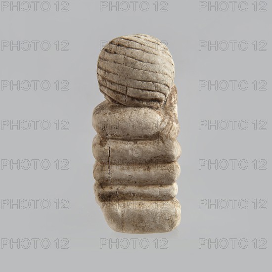 Fragment of a figurine. Head of a polychrome figurine, which originally probably depicted a woman lying on a bed with her head propped up on a pillow. The woman's head is shown wearing a short, striped wig. Behind the head, a woman's hand is visible, which, like the head, rests on an object that may represent a pillow or bolts of cloth. 332 BC–395 AD; Ptolemaic Period–Roman Period. Egypt. Figurine. Height 2.8 cm; width 5.6 cm; depth 2.5 cm., A small sculpted object sits against a plain light gray background, appearing in shades of beige, tan, and light brown with darker brown weathered lines and spots; the top portion is a rounded shape with carved horizontal grooves, and below it a stacked, segmented column of rounded rectangular forms shows visible cracks and textured surface wear, with shadows and subtle highlights accentuating the carved contours and irregularities.