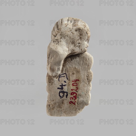 Head of a polychrome figurine, which originally probably depicted a woman lying on a bed with her head propped up on a pillow . The woman's head is shown wearing a short, striped wig. Behind the head, a woman's hand is visible, which, like the head, rests on an object that may represent a pillow or bolts of cloth. Fragment of a figurine. 332 BC–395 AD; Ptolemaic Period–Roman Period. Egypt. Figurine. Height 2.8 cm, width 5.6 cm, depth 2.5 cm., A weathered, pale stone fragment shaped like a small carved figure is centered against a plain light gray background, showing a rounded head with carved hair and a draped section flowing down the side; the stone surface displays a range of off-white, beige, tan, and light brown tones with darker brown weathering and small rough cracks and pits, and a few traces of darker markings on the stone's face and body.