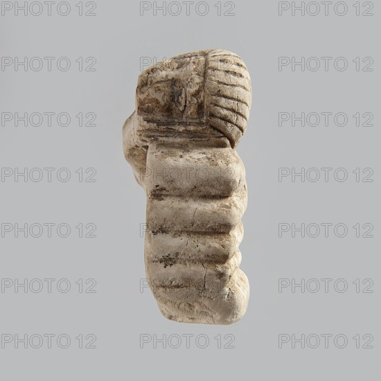 Fragment of a figurine. Head of a polychrome figurine, which originally probably depicted a woman lying on a bed with her head propped up on a pillow . The woman's head is shown wearing a short, striped wig. Behind the head, a woman's hand is visible, which, like the head, rests on an object that may represent a pillow or bolts of cloth. 332 BC–395 AD; Ptolemaic Period–Roman Period. Egypt. Figurine. Height 2.8 cm; width 5.6 cm; depth 2.5 cm., A worn stone fragment with a rounded top and layered, ribbed texture is shown against a plain grey background, displaying beige and light brown tones with darker brown weathered streaks and patches, and visible cracks and surface pitting.