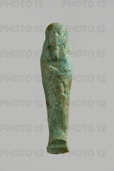 Ushabti figurine, very schematically designed. Distinct narrow beard, tools in crossed hands. Flat back, base under feet. Third Intermediate Period, Egypt; ushabti. Height 10 cm, width 2.5 cm, depth 1.6 cm., A small elongated figurine stands upright against a plain light gray background, with a tapered, shrouded form and a rounded head. The surface is rough and weathered with areas of pitting and encrustation. The object is predominantly green with mottled tones of turquoise and darker green, and scattered patches of brown and tan earth residue. The shape suggests folded arms across the chest and subtle raised features on the head and torso, with a slightly wider base that appears worn. The overall appearance is matte and textured with irregular coloration.