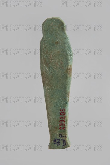 Ushabti, gift, very schematically designed ushabti figurine with a distinct narrow beard and tools in crossed hands; flat back and base under feet. Third Intermediate Period, Egypt; height 10 cm, width 2.5 cm, depth 1.6 cm., A vertical, elongated object with a tapered bottom and a rounded top, showing a rough, pitted surface and irregular edges; its predominant color is a greenish-blue with areas of brownish discoloration and darker speckles, set against a plain light gray background.