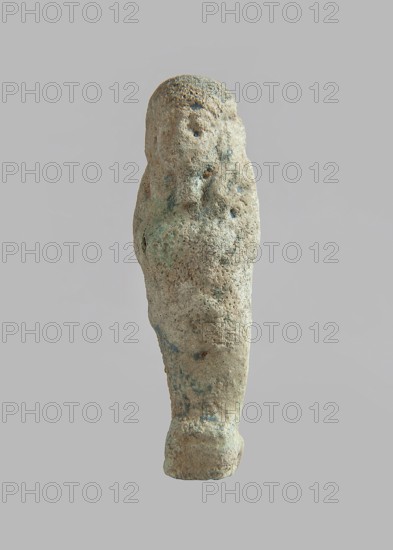 Ushabti, carelessly made figurine with schematic facial features, in a narrow wig revealing the ears, with a rather short beard. In his hands he holds a mattock. The back is flat, the end of the wig is marked with an engraved line. Feet on a pedestal. Glaze almost completely worn away. Third Intermediate Period, Egypt; ushabti; height 6.6 cm, width 2 cm, depth 1.4 cm., A roughly vertical stone object with an irregular, weathered surface displayed against a plain light gray background, showing a textured form that tapers toward the bottom and widens at the top, with subtle rough bumps and pitted areas; the surface color is primarily light beige with patches of pale green and small areas of bluish-gray, as well as darker gray speckles, and the object casts no visible shadow on the background.