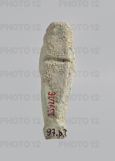 Ushabti. Carelessly made figurine with schematic facial features, in a narrow wig revealing the ears, with a rather short beard. In his hands he holds a mattock. The back is flat; the end of the wig is marked with an engraved line. Feet on a pedestal. Glaze almost completely worn away. Third Intermediate Period, Egypt. Ushabti; height 6.6 cm, width 2 cm, depth 1.4 cm., Gift; author/, A textured vertical object resembling a small carved figure with a rough, pitted surface in shades of beige and light tan with patches of pale green and muted blue, photographed against a plain light gray background; the object tapers slightly toward one end and has subtle ridges and indentations along its length, with a few darker specks and areas of variation in color.