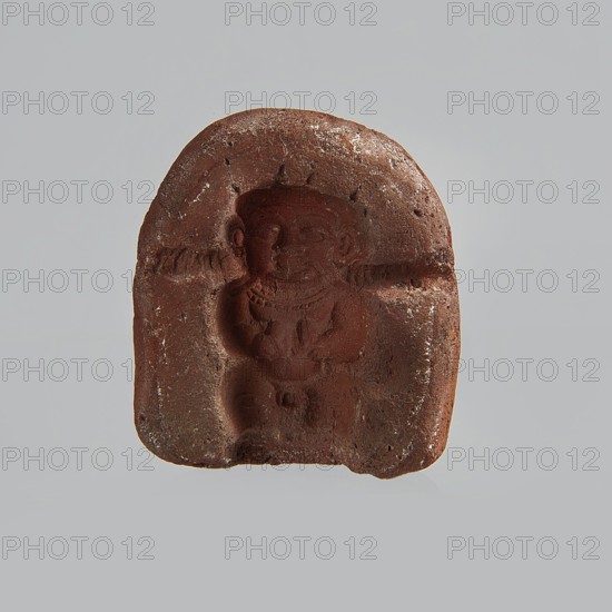 Mold for a figurine of Ptah the dwarf. Mold for pressing casting? a figurine of Ptah the dwarf probably made by pressing from a stone matrix. The head wears a flat cap; a wide face with long, thin mustaches or swallowed snakes; in the hands two oval objects feathers?; legs are curled up. The upper torso is covered by a beaded necklace; otherwise the figure is naked. Technological grooves on both sides of the figurine. Small incisions above the head. The form is semi-oval, rounded at the bottom. Ptah the dwarf mythology; snakes; feathers; necklace iconography. Late Period, Egypt, 664–332 BC. Form. Height 2 cm, width 4.2 cm, depth 4.7 cm., A small reddish-brown carved object with an arched top, showing the recessed figure of a seated person with rounded facial features and a hat or headdress, the figure appearing to wear a necklace and a garment with a defined torso and knees; the surface has a rough, slightly pitted texture with lighter tan and white speckles and chips, and the backgrou...