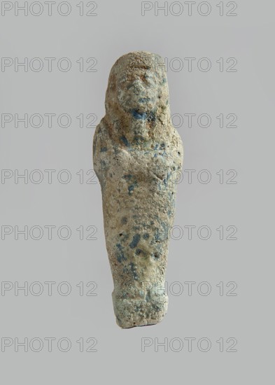Ushabti. Carelessly made figurine with schematic facial features, in a narrow wig revealing the ears, with a rather short beard. In his hands he holds a mattock. The back is flat; the end of the wig is marked with an engraved line. Feet on a pedestal. Glaze almost completely worn away. Third Intermediate Period, Egypt. Ushabti; height 6.5 cm, width 2.1 cm, depth 1.3 cm., A small worn figurine with a tapered, elongated body and a rounded head, showing a rough, pitted surface texture with several irregular spots of darker coloration; the object displays muted beige and tan tones with areas of blue-green and gray mottling, and appears to have faint, shallow contours suggesting facial features and shoulder forms while the lower end is slightly flattened.