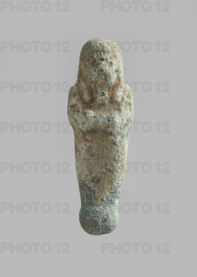 Ushabti. Carelessly made figurine with schematic facial features, in a narrow wig revealing the ears, with a rather short beard. In his hands he holds a mattock. The back is flat; the end of the wig is marked with an engraved line. Feet on a pedestal. Glaze almost completely worn away. Third Intermediate Period, Egypt. Ushabti; height 6.5 cm, width 2 cm, depth 1.4 cm. Maker and exact provenance unknown., A small weathered figurine shaped like a standing human figure with crossed arms, showing a rounded head and stylized facial features, with a narrow, tapered lower body and a rounded base; the surface appears rough and pitted with areas of erosion and encrustation, and the visible colors include pale beige, sandy brown, muted greenish-blue, and touches of darker bluish-gray.