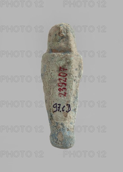 Ushabti. Carelessly made figurine with schematic facial features, in a narrow wig revealing the ears, with a rather short beard. In his hands he holds a mattock. The back is flat; the end of the wig is marked with an engraved line. Feet on a pedestal. Glaze almost completely worn away. Third Intermediate Period, Egypt. Ushabti; height 6.5 cm, width 2 cm, depth 1.4 cm.,  noted., A small, elongated carved figure photographed against a plain light gray background; the object has a rounded top and tapers toward the bottom, with a roughly textured surface showing weathering and small pits; visible colors include pale beige and light tan across most of the surface, patches of bluish-green and gray, and small areas of darker brown; the form suggests a simplified human-like shape with a head-like top and a narrower lower section, and there are a few irregular surface deposits and spots of discoloration.