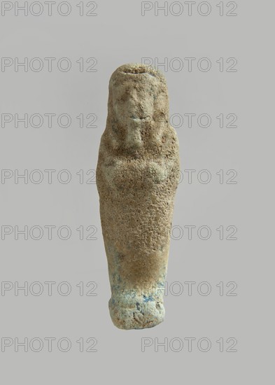 Ushabti. Carelessly made figurine with schematic facial features, in a narrow wig revealing the ears, with a rather short beard. In his hands he holds a mattock. The back is flat, the end of the wig is marked with an engraved line. Feet on a pedestal. Glaze almost completely worn away. Third Intermediate Period, Egypt. Ushabti; height 6.3 cm, width 1.9 cm, depth 1.3 cm., A single upright carved figure with a textured, weathered surface in a range of beige and light brown tones with patches of pale green and faint blue, featuring a stylized head with a rounded top and minimal facial contours and a tapered body that narrows toward the base, set against a neutral gray background.
