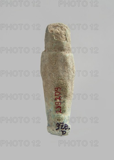 Ushabti. Carelessly made figurine with schematic facial features, in a narrow wig revealing the ears, with a rather short beard. In his hands he holds a mattock. The back is flat, the end of the wig is marked with an engraved line. Feet on a pedestal. Glaze almost completely worn away. Third Intermediate Period, Egypt. Ushabti. Height 6.3 cm, width 1.9 cm, depth 1.3 cm.,  noted., A textured cylindrical object with a rounded top and slightly tapered bottom suspended against a pale gray background, showing a rough, uneven surface with visible pitting and wear; the object displays a mix of beige, light brown, pale green, and hints of bluish-green patina across its surface, and a small area near the base appears darker with muted blackish markings.