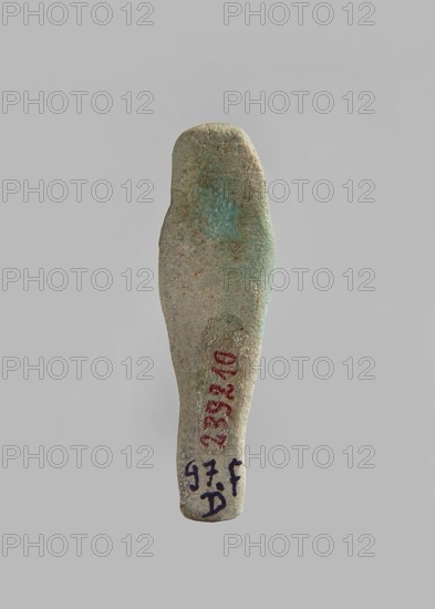 Ushabti, Mass-produced ushabti figurine with very schematic shapes, with a barely marked face and crossed arms. Flat back. Glaze partially discolored. Third Intermediate Period, Egypt. Ushabti; height 5.8 cm, width 1.7 cm, depth 1 cm., A single elongated object shaped like a stylized human figure is centered against a plain light gray background; the object has a rounded top and tapers toward the bottom, with a slightly uneven, worn surface showing a mix of muted green, pale turquoise, beige, and gray tones, plus areas of brownish discoloration and small rough textures; there are traces of darker markings near the lower portion in a dark bluish-black and some faded red, and the object casts a faint shadow on the background.