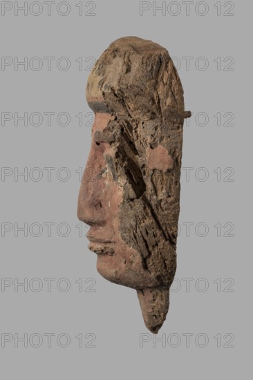 Sarcophagus mask with a partially preserved fragment of a wig above the forehead painted yellow with traces of black polychrome. It is separated from the forehead by cherry-red and black bands. The eyes are relatively large, with black rims, pupils, and eyebrows; the whites are white. The nose is long, quite narrow, widening evenly towards the nostrils. The mouth is primitively modeled. A fragment of the neck is preserved under the chin. The face is painted cherry-red. Partially preserved mounting pegs at the back. 332–30 BC; Ptolemaic Period, Egypt. Sarcophagus mask; height 25.7 cm, width 18 cm, thickness 8.2 cm., A weathered carved wooden profile of a human face with a pronounced nose and lips, shown against a plain gray background; the surface is rough and eroded with visible cracks and holes, and patches of faded pink and reddish pigment mixed with natural brown and beige wood tones, along with some darker gray-brown areas.